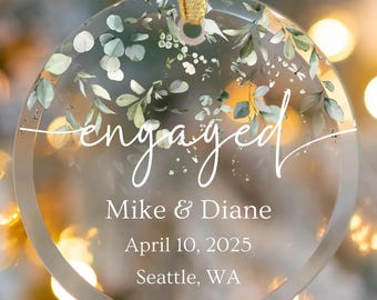 May include: A clear, round ornament with the word "engaged" in elegant script. The ornament features floral accents and the names "Mike & Diane," along with the date "April 10, 2025," and "Seattle, WA."
