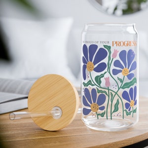 K&ouml;nnte beinhalten: Eine Glaswasserflasche mit Bambusdeckel und Strohhalm. Die Flasche hat ein Blumenmuster mit blauen Blumen und gr&uuml;nen Bl&auml;ttern. Der Text "BE PROUD OF YOUR PROGRESS" ist auf die Flasche gedruckt.