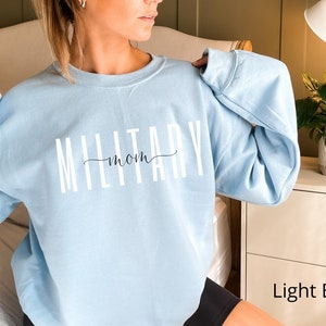 Puede incluir: Sudadera azul claro con la frase "MILITARY mom" en letras blancas y una "mom" en cursiva. La sudadera tiene mangas largas y cuello redondo. Las palabras est&aacute;n centradas en el pecho.