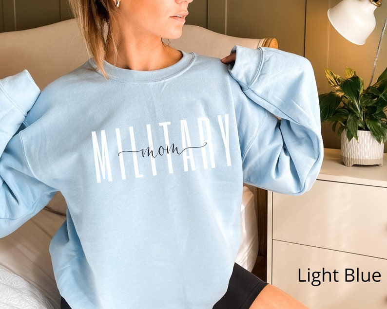 Puede incluir: Sudadera azul claro con la frase "MILITARY mom" en letras blancas y una "mom" en cursiva. La sudadera tiene mangas largas y cuello redondo. Las palabras est&aacute;n centradas en el pecho.
