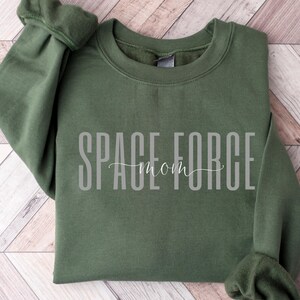 Puede incluir: Una sudadera verde oscuro con el texto "SPACE FORCE mom" en letras blancas.