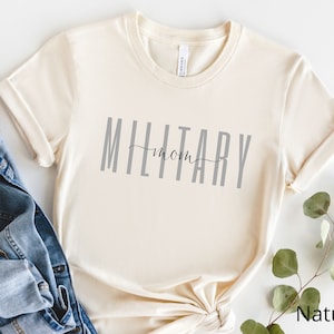 Pode incluir: Uma camiseta de cor natural com o texto "MILITARY mom" impresso em cinza na frente.