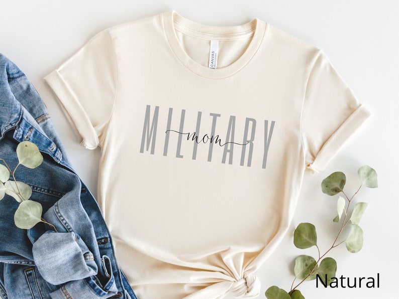 Pode incluir: Uma camiseta de cor natural com o texto "MILITARY mom" impresso em cinza na frente.