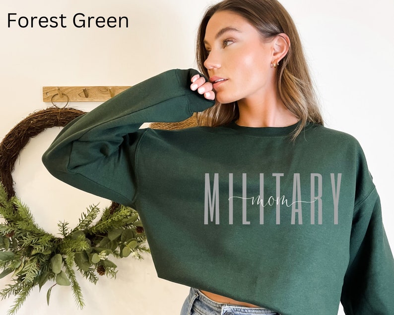 Puede incluir: Sudadera verde bosque con el texto "MILITARY mom" en gris. La sudadera es de color liso y tiene cuello redondo. El texto est&aacute; en una fuente sans-serif con la palabra "mom" en una fuente cursiva.