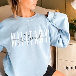 K&ouml;nnte beinhalten: Hellblauer Pullover mit dem Text "Military girlfriend" in wei&szlig;en Buchstaben.
