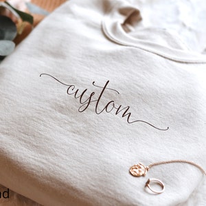 May include: A sand-colored sweatshirt with the word "custom" written in cursive on the front.