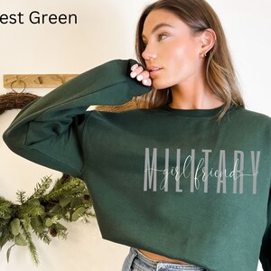 K&ouml;nnte beinhalten: Gr&uuml;nfarbener, kurzer Sweatshirt mit dem Text "Military Girlfriend" in grauer Schrift.