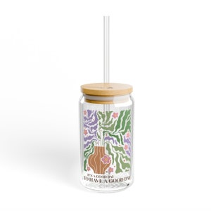 K&ouml;nnte beinhalten: Ein durchsichtiger Glasbecher mit Bambusdeckel und Strohhalm. Der Becher hat ein florales Design mit einer Vase und dem Text "It's a good day to have a good day".