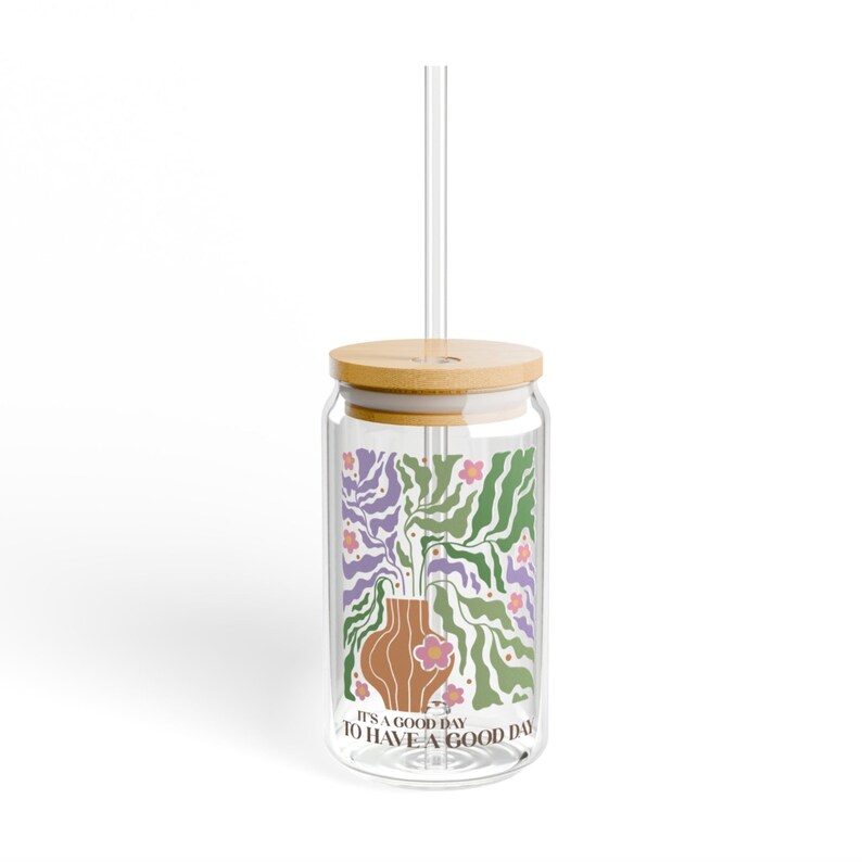 K&ouml;nnte beinhalten: Ein durchsichtiger Glasbecher mit Bambusdeckel und Strohhalm. Der Becher hat ein florales Design mit einer Vase und dem Text "It's a good day to have a good day".