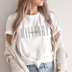Pode incluir: Camiseta branca com o texto "MILITARY mom" em letras cinzas.