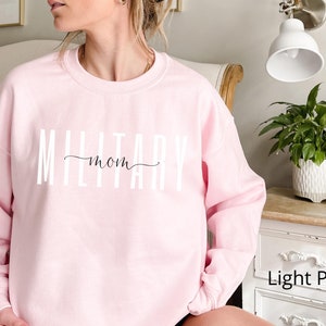 Puede incluir: Sudadera rosa claro con el texto "MILITARY mom" en letras blancas y cursiva. La sudadera es de color rosa claro y tiene cuello redondo. Las palabras "Light Pink" est&aacute;n en la esquina inferior derecha.