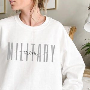 Puede incluir: Sudadera blanca con el texto "MILITARY mom" en letras grises y "mom" en cursiva. La imagen es una toma media de la parte superior del cuerpo. La sudadera es de estilo casual.