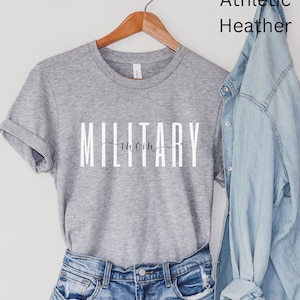 Pode incluir: Uma camiseta cinza com o texto "MILITARY mom" em letras brancas. A camiseta est&aacute; pendurada em um cabide de madeira e &eacute; exibida em um fundo branco.