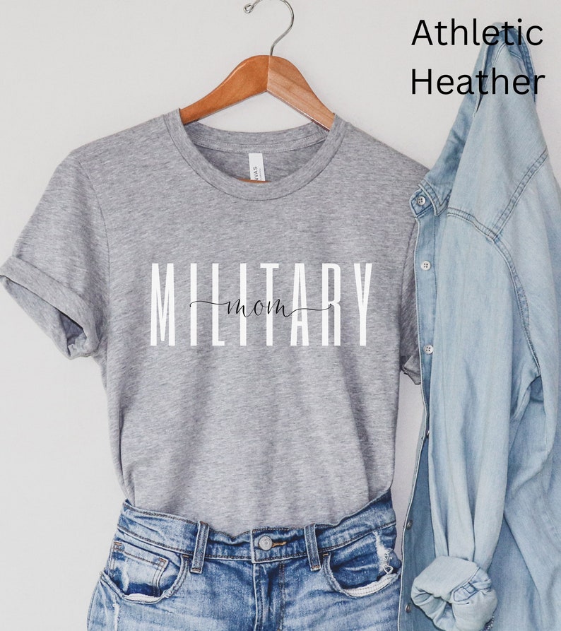 Pode incluir: Uma camiseta cinza com o texto "MILITARY mom" em letras brancas. A camiseta est&aacute; pendurada em um cabide de madeira e &eacute; exibida em um fundo branco.