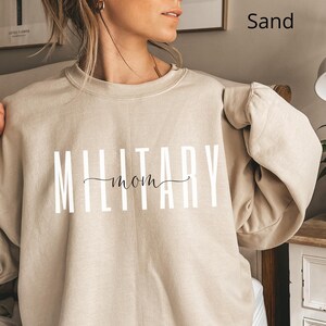 Puede incluir: Sudadera color arena con el texto "MILITARY mom" en letras blancas y un "mom" en cursiva. La sudadera de manga larga es de color neutro.