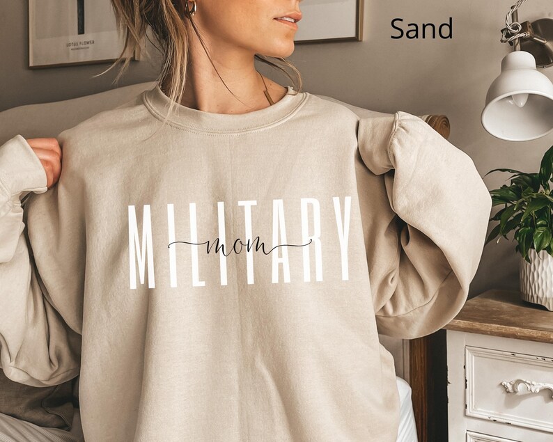 Puede incluir: Sudadera color arena con el texto "MILITARY mom" en letras blancas y un "mom" en cursiva. La sudadera de manga larga es de color neutro.