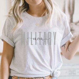 Pode incluir: Uma camiseta branca com o texto "Military Mom" impresso em letras cinzas. A camiseta est&aacute; sendo usada por uma pessoa com cabelo loiro.