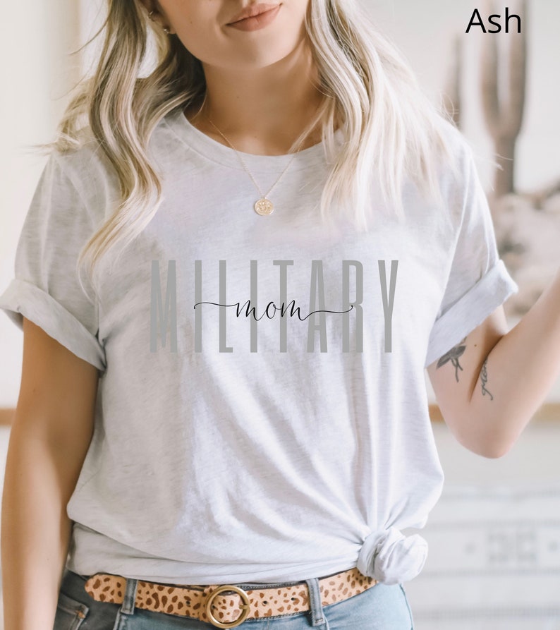 Pode incluir: Uma camiseta branca com o texto "Military Mom" impresso em letras cinzas. A camiseta est&aacute; sendo usada por uma pessoa com cabelo loiro.