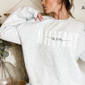 Puede incluir: Sudadera gris ceniza con el texto "MILITARY mom" en letras blancas y un "mom" en cursiva. La sudadera de cuello redondo tiene mangas largas. El fondo es neutro.