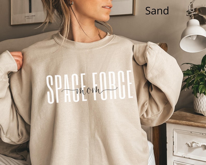 Puede incluir: Una sudadera de color arena con el texto "SPACE FORCE mom" impreso en blanco en la parte delantera.