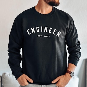 May include: Black crew neck sweatshirt with the word "ENGINEER" arched across the chest in white, with "EST. 2022" below. The sweatshirt is a solid colour and has long sleeves. The overall theme is apparel.