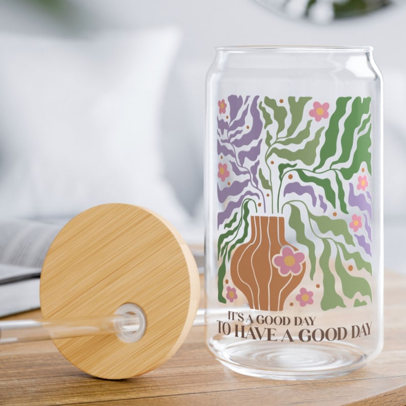 K&ouml;nnte beinhalten: Eine Glaswasserflasche mit Bambusdeckel und einem farbenfrohen Blumendesign. Auf der Flasche steht der Text "It's a good day to have a good day".