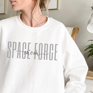 Puede incluir: Sudadera blanca con el texto "SPACE FORCE mom" impreso en gris en la parte delantera.