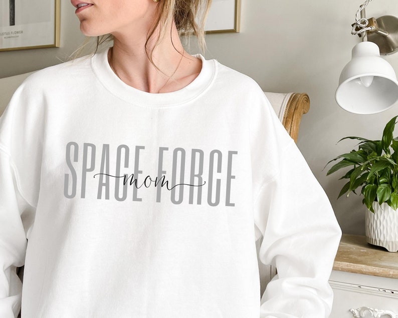 Puede incluir: Sudadera blanca con el texto "SPACE FORCE mom" impreso en gris en la parte delantera.