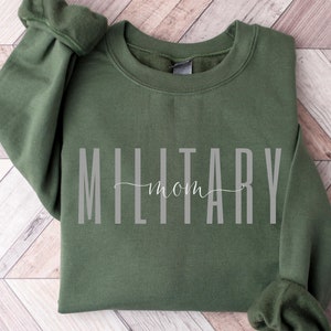 Puede incluir: Una sudadera verde oliva con las palabras "MILITARY mom" impresas en gris. El texto est&aacute; centrado en la parte delantera de la prenda. La sudadera est&aacute; doblada sobre una superficie de madera marr&oacute;n claro.