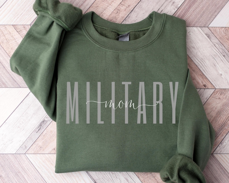 Puede incluir: Una sudadera verde oliva con las palabras "MILITARY mom" impresas en gris. El texto est&aacute; centrado en la parte delantera de la prenda. La sudadera est&aacute; doblada sobre una superficie de madera marr&oacute;n claro.
