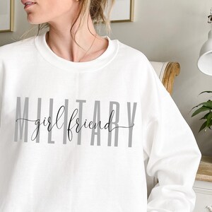 K&ouml;nnte beinhalten: Wei&szlig;er Pullover mit dem Aufdruck "MILITARY girlfriend" in Grau auf der Vorderseite.
