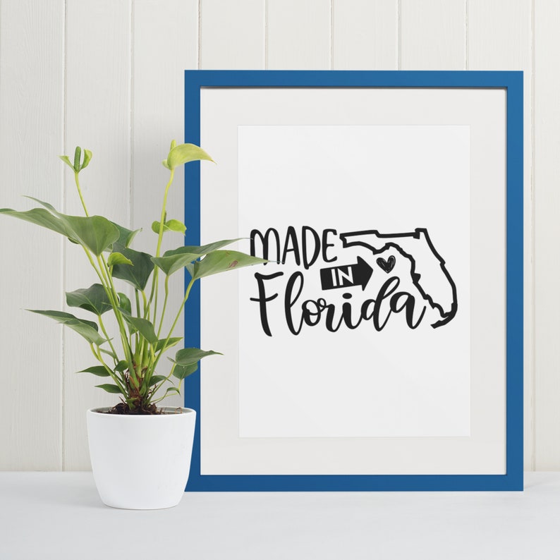 May include: A framed print with a blue border and white matting. The print features the words "MADE IN Florida" in a black, cursive font, with a black outline of the state and a heart. A potted plant sits to the left.