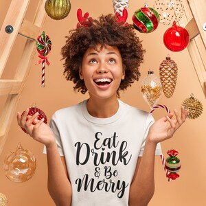 May include: A white t-shirt with the text "Eat Drink & Be Merry" in black lettering. The person wearing the shirt is surrounded by Christmas ornaments, including candy canes, pinecones, and baubles.