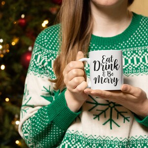 May include: White ceramic mug with black text that reads "Eat Drink & Be Merry". A person is holding the mug in front of a Christmas tree.