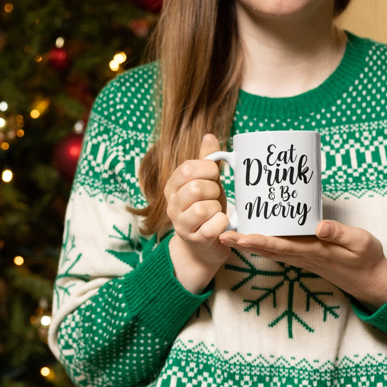 May include: White ceramic mug with black text that reads "Eat Drink & Be Merry". A person is holding the mug in front of a Christmas tree.