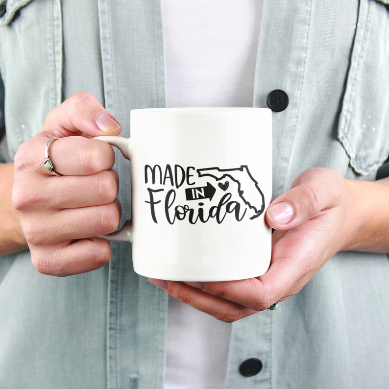 May include: A white ceramic mug with the words "MADE IN Florida" in black script and a black outline of the state. The mug is held by a person wearing a light green jacket.