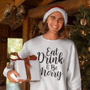 May include: A white Christmas sweater with the text "Eat Drink & Be Merry" in black script. The person wearing the sweater is holding a white gift box with a red and green ribbon.