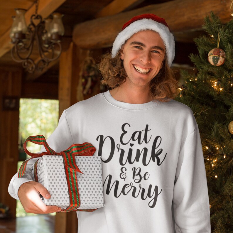 May include: A white Christmas sweater with the text "Eat Drink & Be Merry" in black script. The person wearing the sweater is holding a white gift box with a red and green ribbon.