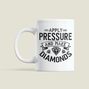 Peut inclure: Une tasse en c&eacute;ramique blanche avec un motif noir qui dit "=APPLY= PRESSURE AND MAKE DIAMONDS" avec trois diamants en dessous du texte.