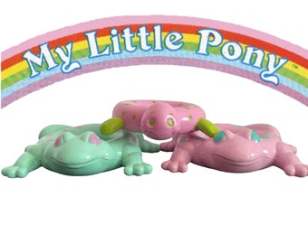 May include: Three My Little Pony frog figurines. One is green with pink spots, one is pink with green spots, and one is a pink and green inflatable ring.