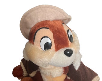 Puede incluir: Un peluche de un personaje de dibujos animados, una ardilla, con colores marrón, beige y crema. La ardilla lleva una chaqueta marrón con ribetes beige y una gorra beige. El juguete tiene grandes ojos negros y una nariz negra.
