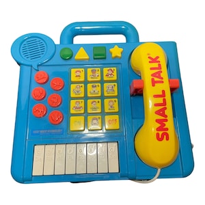 May include: A blue toy phone with a yellow receiver and a red button. The phone has a keypad with numbers and pictures of people, a small piano keyboard, and the words "SMALL TALK" on the side.