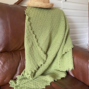 Puede incluir: Una manta de crochet verde con un borde festoneado está colocada sobre un sillón de cuero marrón. Un sombrero de paja está colocado sobre la manta.