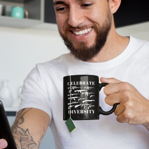 May include: A black coffee mug with a white design that says "Celebrate Diversity" and features a row of different types of guns. The mug is being held by a person with a tattoo on their arm.