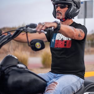 Może przedstawiać: Mężczyzna w czarnym t-shircie z nadrukiem flagi Teksasu i napisem "Born & Bred". Jedzie na motocyklu.