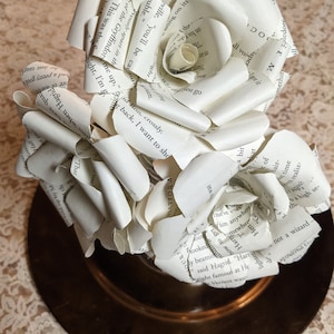 Pode incluir: Um buquê de três rosas brancas de papel feitas com as páginas de um livro. As rosas estão dispostas em um prato marrom com uma toalha de renda branca por baixo.