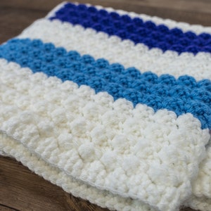 Puede incluir: Una manta blanca de ganchillo con rayas azules, rojas y rosas. La manta está hecha con un punto texturizado que crea una superficie abultada.
