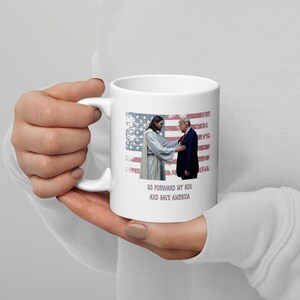 May include: White ceramic coffee mug with a graphic of Jesus laying his hand on Donald Trump's shoulder. The graphic is set against a red, white, and blue American flag background. The text "Go forward my son and save America" is printed below the graphic.