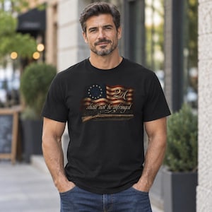 May include: A black short-sleeved t-shirt featuring a graphic of a distressed American flag with a circle of thirteen stars, the text 2A, and the phrase shall not be infringed, accompanied by an illustration of a long rifle at the bottom.