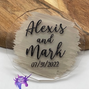 May include: A clear acrylic sign with a beige brushstroke design. The text "Alexis and Mark 07/31/2022" is printed in black.